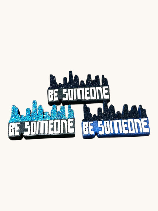 Flutterby Creations car freshie with "BE SOMEONE" text and a glittery black skyline design. Available in blue and black.