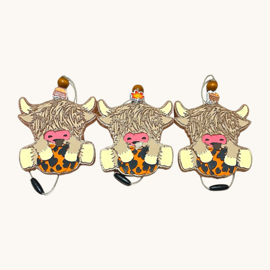 Flutterby Creations Highland cow car freshie with pumpkin design, featuring fluffy brown fur, pink nose, and orange/black spotted body.